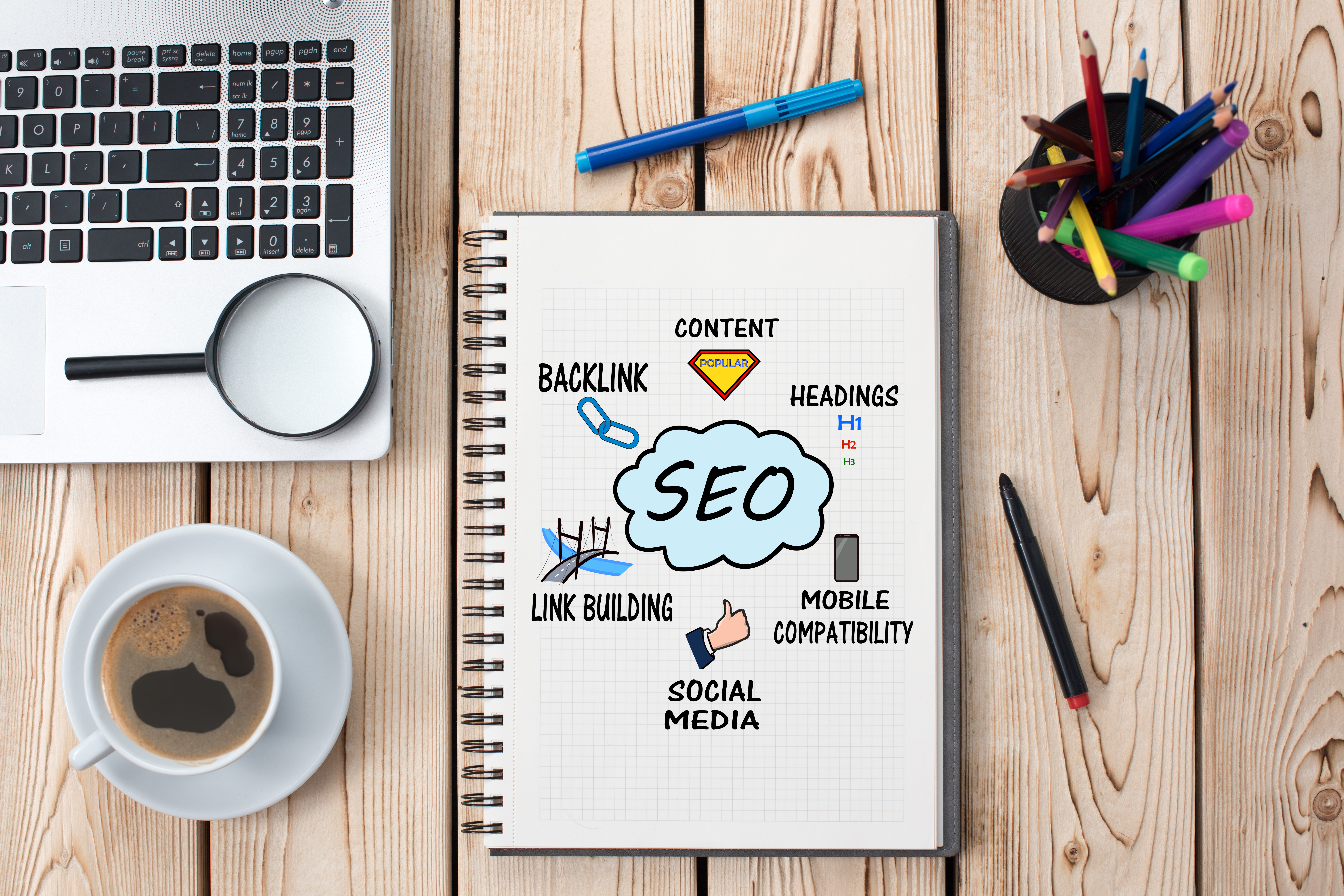 a notebook with SEO written in the middle of it, on a table with a laptop, a cup of coffee, and several coloured pens